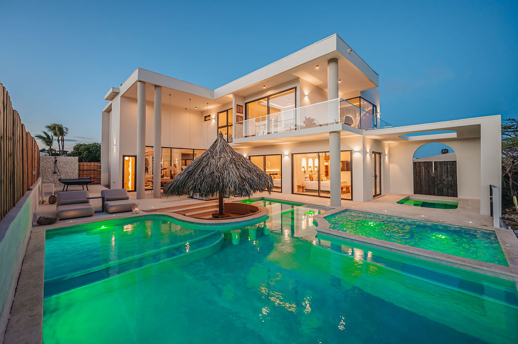 A Mediterranean-inspired villa at twilight, glowing with turquoise pool light and a thatched palapa